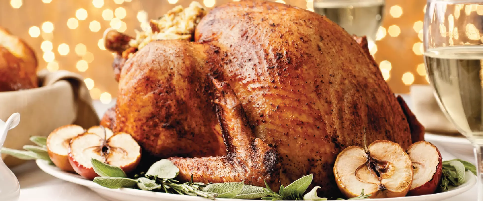 A large turkey on a plate with sage and dried apples. Wine glasses and bread are also on the table. Lights are in the background..